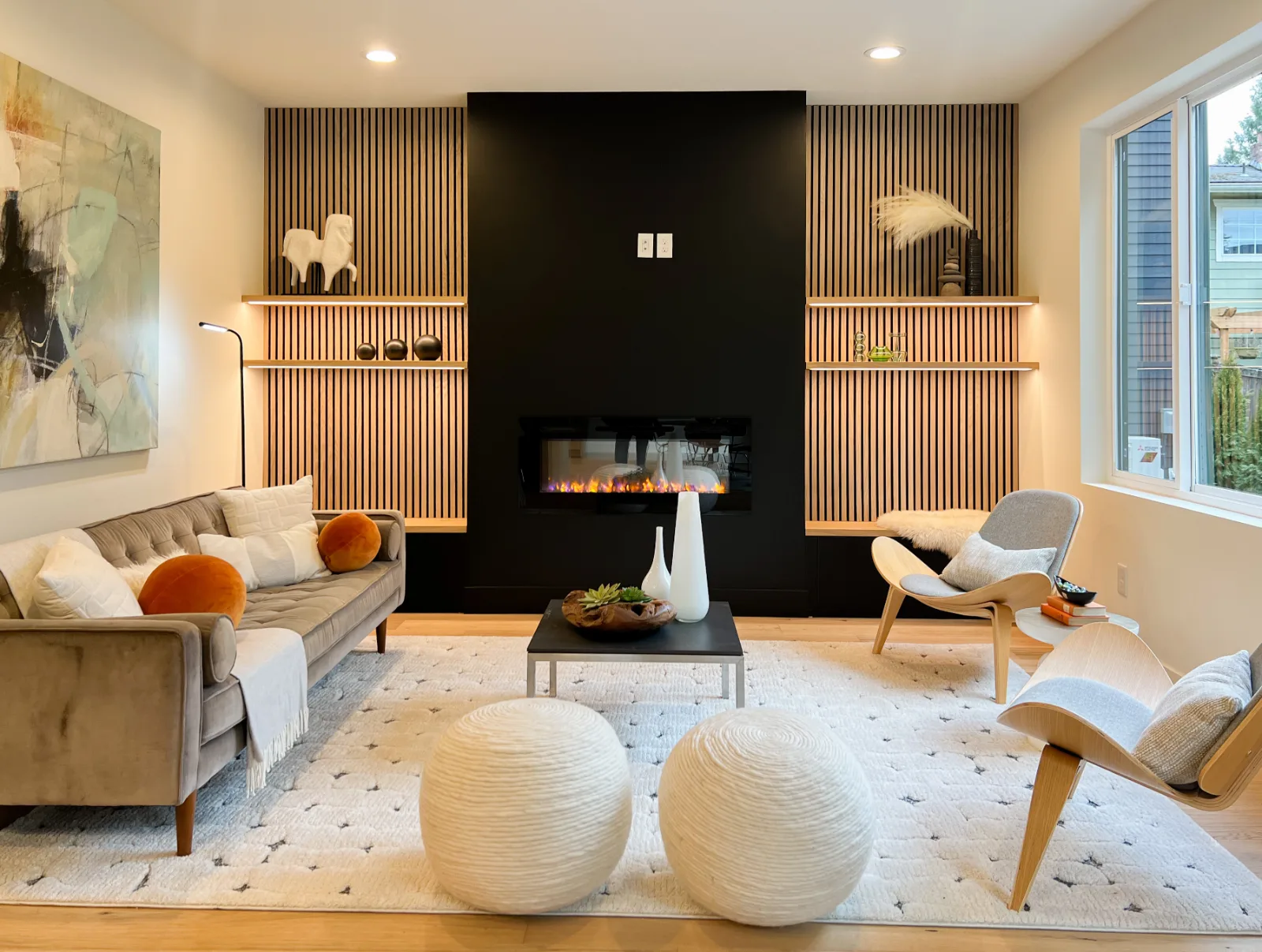 A modern mid-century living room features a sleek black fireplace flanked by slatted wood accent walls, a velvet sofa, two bentwood accent chairs, and round white poufs.
