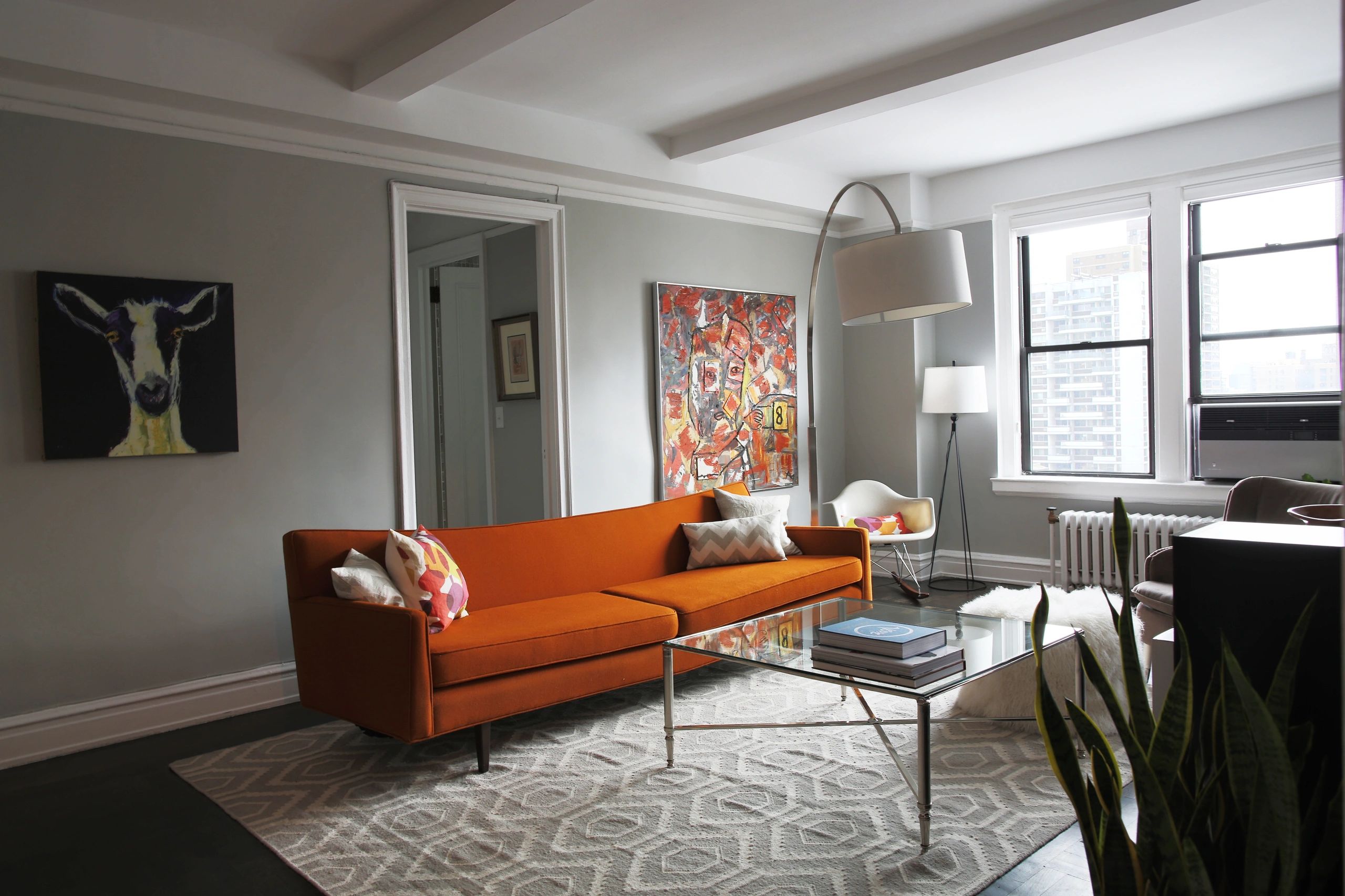 A stylish mid-century modern living room features a vibrant orange sofa, an glass coffee table on a patterned rug, and abstract art pieces against light grey walls.