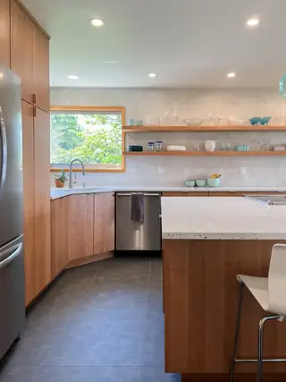 A modern kitchen features warm wood cabinetry, open floating shelves, white speckled quartz countertops, and dark gray tiled floors.