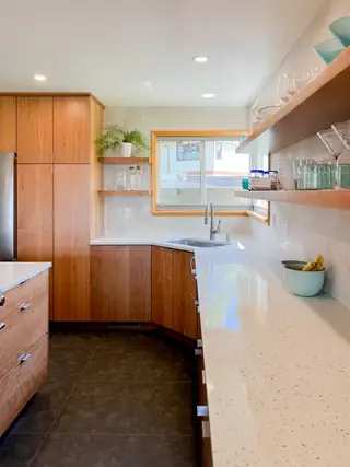 A modern kitchen features warm wood cabinetry, terrazzo countertops, floating open shelves, and dark tile flooring.