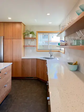 A bright contemporary kitchen featuring warm wooden cabinetry, white terrazzo countertops, open floating shelves, and a corner sink.