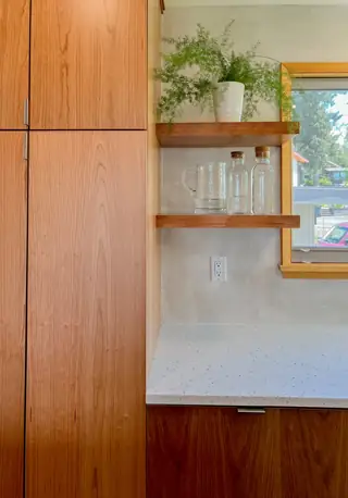 A modern kitchen corner features natural wood cabinetry, floating shelves with glass carafes, a potted fern, and a white terrazzo countertop.