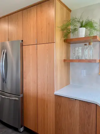 A modern kitchen corner features warm wood cabinetry, a stainless steel refrigerator, and floating shelves mounted above a light speckled countertop.