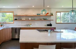 A bright modern kitchen features open wooden floating shelves, turquoise glass pendant lights, and a large white terrazzo island.