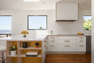 Blue Ridge Remodel: A modern white kitchen features a terrazzo-topped island with mustard yellow open shelving, light wood accents, and a minimal range hood