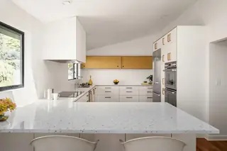 Blue Ridge Remodel: A bright mid-century modern kitchen features speckled white terrazzo countertops, mustard yellow upper cabinets, and integrated stainless steel appliances