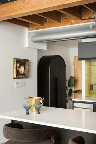 Capitol Hill Loft: A modern industrial kitchen features a white quartz island counter with barstools, an arched black cabinet, and exposed wooden ceiling beams