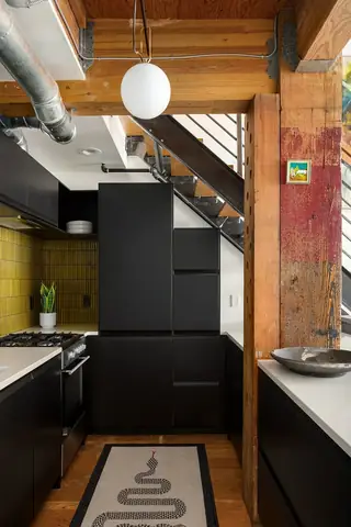 Capitol Hill Loft: A modern industrial loft kitchen features sleek black cabinetry, a yellow tiled backsplash, exposed wooden beams, and a snake-patterned runner rug