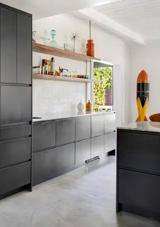 A modern kitchen features sleek dark charcoal cabinetry, white marble countertops, floating wooden shelves against a white brick wall, and a colorful retro rocket statue in the corner.