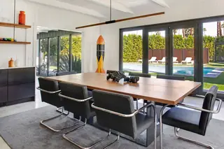 A mid-century modern dining room features a long wooden table surrounded by black cantilever chairs, set against glass doors overlooking a poolside backyard.