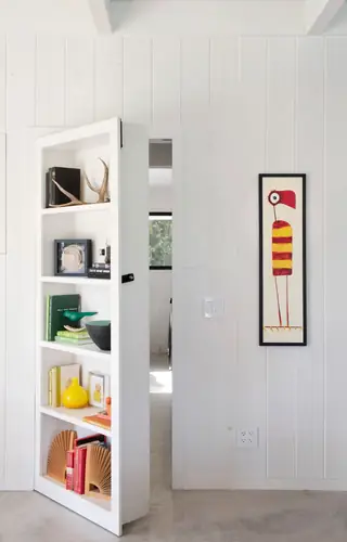 A white built-in bookshelf secret door swings open to reveal an adjoining room inside a modern white-paneled home.