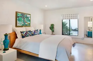 A mid-century modern bedroom featuring a light wood platform bed with white bedding, turquoise accent pillows, and vibrant teal ceramic lamps.