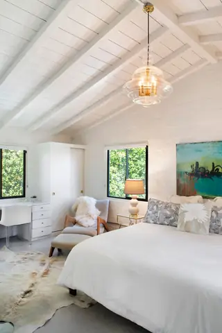 A bright bedroom features white-painted exposed ceiling beams, a glass pendant light, a white duvet on the bed, and a mid-century style armchair with an ottoman.