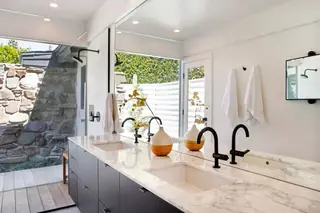A modern bathroom features a double marble vanity with matte black gooseneck faucets and a large wall mirror overlooking an outdoor stone shower enclosure.