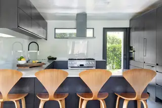 A modern kitchen features a dark gray island lined with four bentwood counter stools set against matching dark cabinetry and a stainless steel range hood.