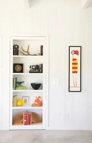 A white built-in bookcase filled with colorful curated decor sits set into a white paneled wall beside a narrow framed bird artwork.