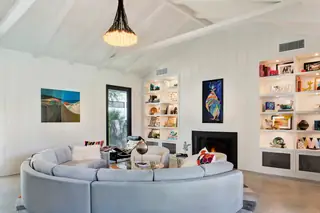 A bright living room features a large light grey curved sectional sofa, vaulted white ceilings with exposed beams, and built-in lit bookshelves flanking a black fireplace.