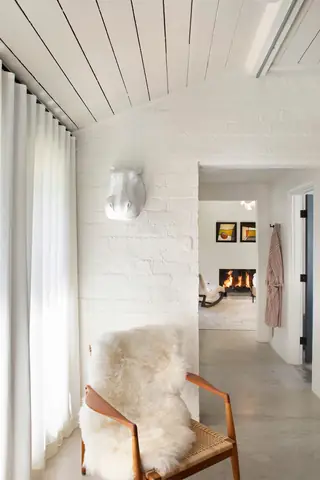 A bright modern room corner features a wooden accent chair draped with a plush sheepskin rug beside a white painted brick wall and a doorway looking through to a fireplace.