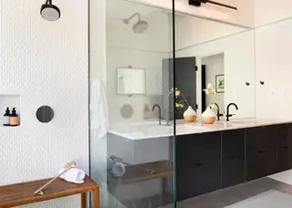 A modern bathroom features a walk-in glass shower with textured white tile, a dark floating vanity, black matte fixtures, and a wooden bench.