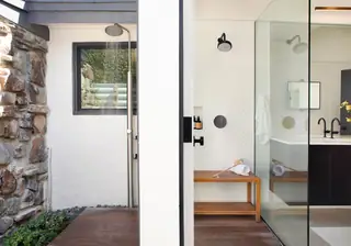 An open doorway highlights a seamless transition between a modern indoor bathroom with a wooden bench and an outdoor shower with a stone wall.