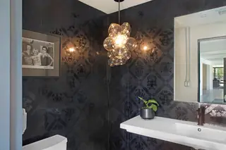 A moody powder room features black damask wallpaper, a dramatic bubble glass pendant light, and a sleek white floating sink.
