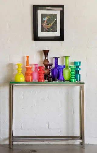 A spectrum of colorful glass vases lines a glass-top metal console table against a white brick wall beneath a framed art print.