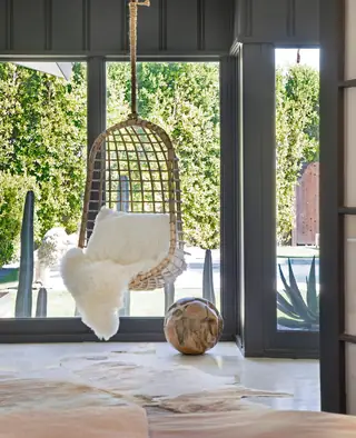 A light-filled modern room features a hanging rattan swing chair draped with sheepskin in front of floor-to-ceiling dark charcoal windows.