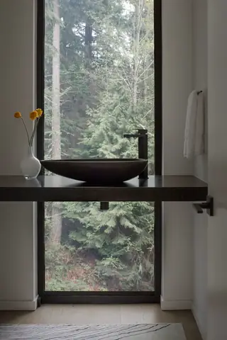 Chuckanut New Build, Bellingham: A modern floating bathroom vanity featuring a dark vessel sink and black faucet stands in front of a floor-to-ceiling window looking onto a lush