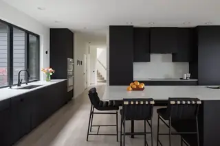 Chuckanut New Build, Bellingham: A modern kitchen features matte black cabinetry, white quartz countertops, light wood flooring, and black bar stools at a spacious island