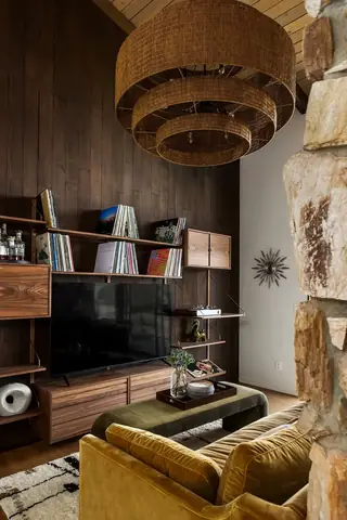 Edmonds Beach House: A mid-century modern living room features dark wood paneling with custom vinyl record shelving, a yellow velvet sofa, and a multi-tiered woven pendant light