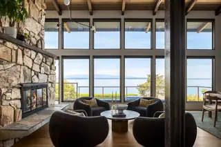 Edmonds Beach House: A cozy modern living room features a grand stone fireplace and dark rounded armchairs arranged around a coffee table facing floor-to-ceiling windows