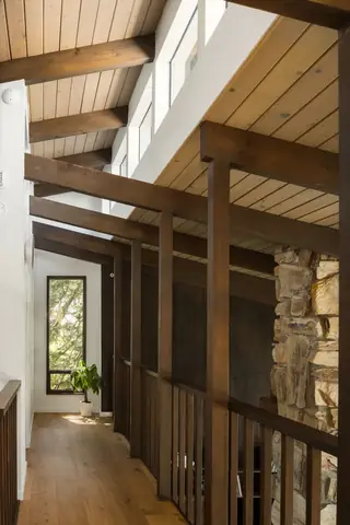Edmonds Beach House: A mid-century modern upper-level hallway features exposed dark wood beams, vaulted plank ceilings, a stone fireplace column, and a potted plant at the far end