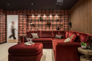 Edmonds Beach House: A moody, retro-inspired lounge features a plush red velvet sectional sofa, matching ottoman, geometric patterned wallpaper, and recessed display shelves