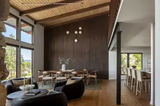 Edmonds Beach House: A mid-century modern open-concept space featuring a dark wood accent wall, vaulted beam ceiling, dark round armchairs, and an adjacent dining area