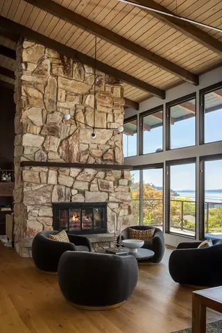 Edmonds Beach House: A mid-century modern living room features a soaring stone fireplace, dark rounded accent chairs, and expansive windows offering scenic coastal views