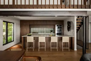 Edmonds Beach House: A modern kitchen features rich walnut cabinetry, a long island with four beige bar stools, and a green tiled backsplash beneath an open loft area