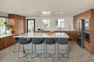 Greenwood Kitchen Design: A spacious modern kitchen featuring rich walnut cabinetry, a large grey island topped with white quartz, navy blue barstools, and terrazzo tile flooring