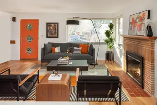 Greenwood Kitchen Design: A mid-century modern living room featuring an orange front door, brick fireplace, dark gray sofa, and low-profile black armchairs on a patterned area rug