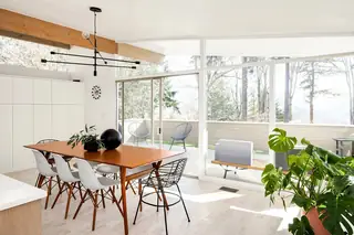 Issaquah Interior Design: A sunlit mid-century modern dining room features a wooden table surrounded by white chairs beneath large glass windows with forest views