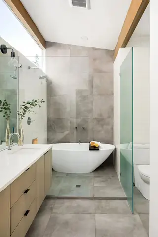 Issaquah Interior Design: A modern bathroom features a white freestanding bathtub set against large grey wall tiles, alongside a light wood vanity and glass partition