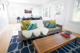 Madison Valley Design: A bright mid-century modern living room features a grey sofa accented with colorful pillows and a faux fur throw, set above a patterned navy rug
