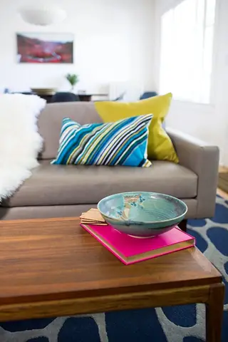 Madison Valley Design: A mid-century modern wooden coffee table features a bright pink book and ceramic bowl in front of a grey sofa with colorful accent pillows