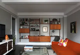 A mid-century modern living room features a teak wall shelving unit, a vibrant orange sofa with a patterned pillow, and a glass coffee table over dark wood floors.