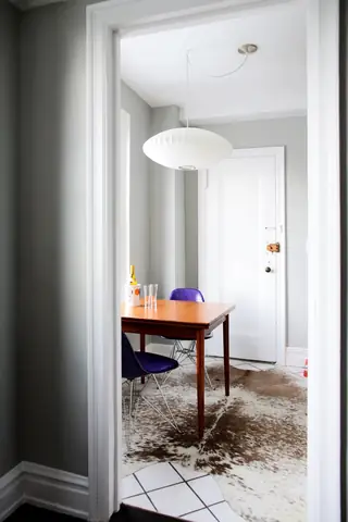 A compact mid-century modern dining area framed by a doorway features a wooden table paired with purple shell chairs, a white saucer pendant light, and a cowhide rug.