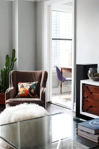 A cozy living room scene features a plush brown velvet armchair with a floral throw pillow, a glass coffee table, and a view into an adjacent sunlit balcony room through an open doorway.