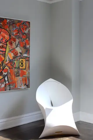A white sculptural chair sits in the corner of a minimalist room beside a large, vibrant abstract painting hanging on a light grey wall.