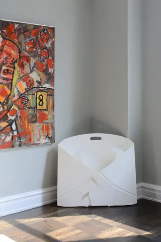 A minimalist room corner displays a unique white sculptural chair beside a vibrant abstract painting on a light gray wall.