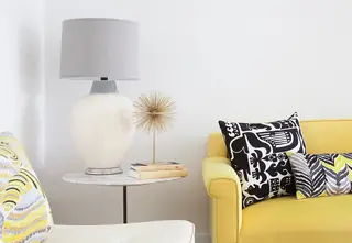 Pebble Beach Drive, Palm Springs: A vibrant living room nook featuring a bright yellow sofa with graphic pillows beside a marble top side table holding a ceramic lamp