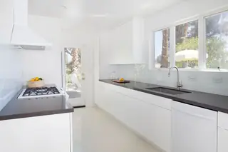Pebble Beach Drive, Palm Springs: A bright modern kitchen features minimalist white cabinetry, dark grey countertops, and a large window overlooking an outdoor yard
