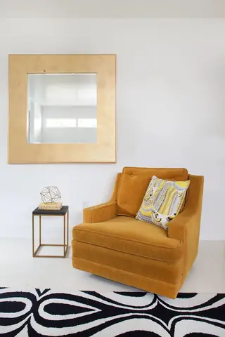 Pebble Beach Drive, Palm Springs: A mustard yellow upholstered armchair with a patterned accent pillow sits next to a small brass side table below a large gold-framed mirror